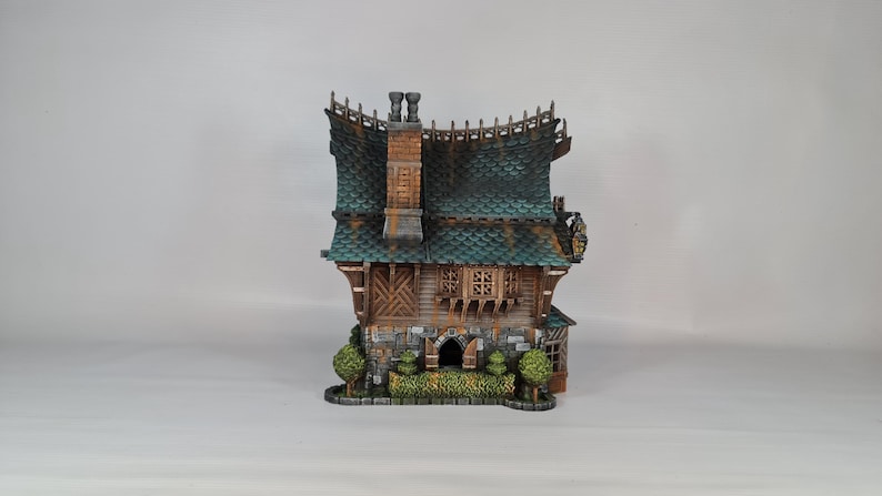 May include: A detailed miniature house with a green tiled roof, wooden siding, and stone accents. The house features a chimney, small windows, and decorative trim. Two small green bushes flank the front of the house.