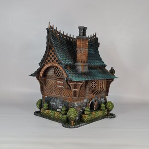 May include: A detailed miniature house with a teal roof, brown wooden walls, and a stone base. The house features a chimney, decorative trim, and small bushes around the base. The overall design has a fantasy or fairy tale aesthetic.