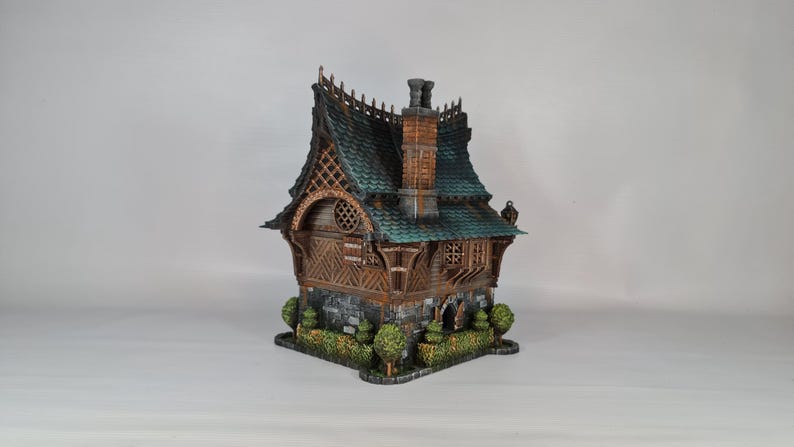 May include: A detailed miniature house with a teal roof, brown wooden walls, and a stone base. The house features a chimney, decorative trim, and small bushes around the base. The overall design has a fantasy or fairy tale aesthetic.