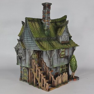 A miniature fantasy house model, with a green roof, gray siding, and stone foundation, is viewed from the front-right, showing a prominent exterior staircase and a small fence.