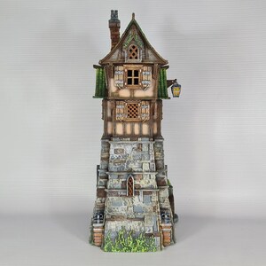 May include: A miniature stone tower with a wooden house on top. The house has a brown roof, green trim, and two windows. The tower has a stone wall with a small arched doorway. The tower is painted in shades of gray and brown, with green accents.