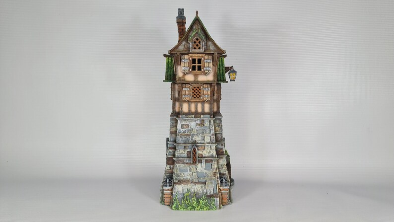 May include: A miniature stone tower with a wooden house on top. The house has a brown roof, green trim, and two windows. The tower has a stone wall with a small arched doorway. The tower is painted in shades of gray and brown, with green accents.
