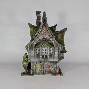 A miniature fantasy house model, with a green roof, gray siding, and stone foundation, is viewed from the front, showing a small green tree and a front door with a window.
