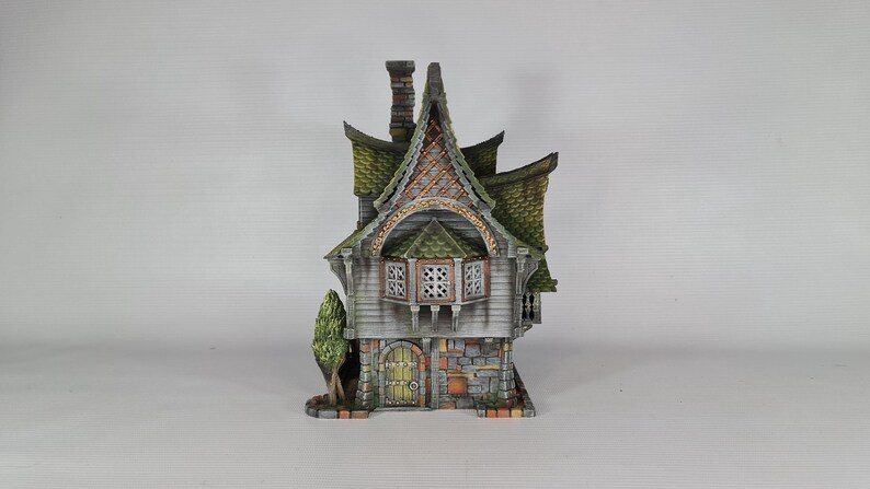 A miniature fantasy house model, with a green roof, gray siding, and stone foundation, is viewed from the front, showing a small green tree and a front door with a window.