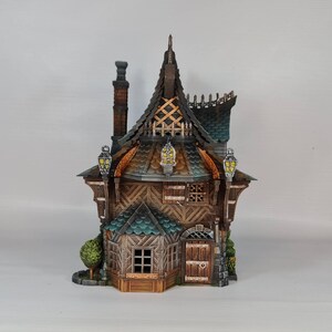 May include: A detailed miniature house with a steeply pitched roof, chimney, and decorative lanterns. The house features a wooden facade with intricate details, including a small front door and windows. The roof is a mix of green and brown shingles.