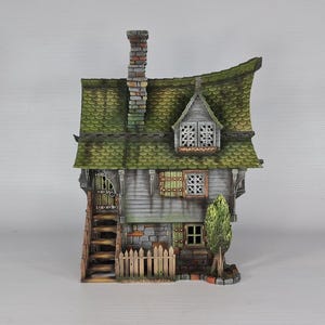A miniature fantasy house model, with a green roof, gray siding, and stone foundation, is viewed from the front, showing an exterior staircase, a small fence, and a green tree.