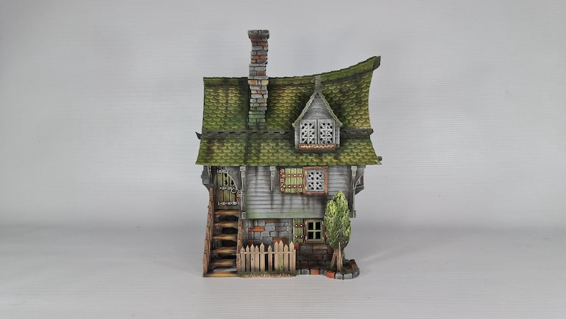 A miniature fantasy house model, with a green roof, gray siding, and stone foundation, is viewed from the front, showing an exterior staircase, a small fence, and a green tree.
