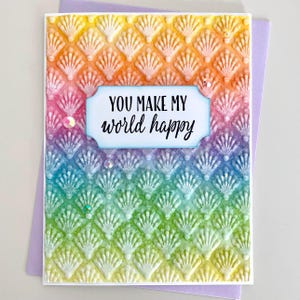 May include: A handmade greeting card with a rainbow gradient background and a seashell pattern. The card features the text "YOU MAKE MY world happy" in black script within a light blue frame. A purple envelope is visible.