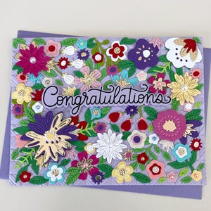 May include: A rectangular greeting card with a light purple embossed background featuring a dense arrangement of colorful paper flowers in shades of pink, purple, yellow, red, and white. The word Congratulations is written in black script.
