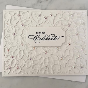 May include: A white rectangular greeting card featuring an intricate laser-cut floral pattern with embedded iridescent rhinestones. A central white label displays the black printed text "TIME TO Celebrate" in a mix of block and script fonts.