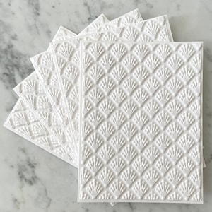 May include: A stack of white, textured cards with a repeating fan-shaped pattern. The cards are arranged in a fanned-out display, showcasing the embossed design. The cards are rectangular and have a clean, modern aesthetic.