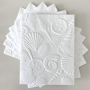 May include: White embossed stationery set featuring various seashell designs. The set includes several rectangular cards with detailed relief patterns of different shells, including conch shells and scallops. The cards are stacked, showcasing the intricate textures and designs.