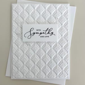 May include: A white greeting card featuring an embossed shell pattern across the entire surface. A rectangular white label is centered on the card with the black printed text "WITH SYMPATHY AND LOVE" in a mix of script and sans-serif fonts.