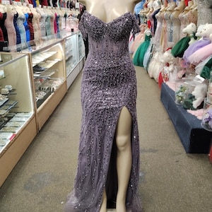 May include: A lavender-colored formal gown with a beaded bodice and a high slit. The dress is displayed on a mannequin in a retail setting, surrounded by other dresses. The gown features off-the-shoulder sleeves and a flowing skirt.