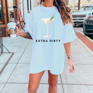 May include: White t-shirt with a martini glass graphic and the text "EXTRA DIRTY". The martini glass has a green olive garnish. The shirt is a casual, oversized style.