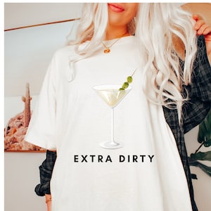 May include: White t-shirt with a martini glass graphic and the text "EXTRA DIRTY". The martini glass has a clear liquid and three green olives on a toothpick. The shirt is a casual style.