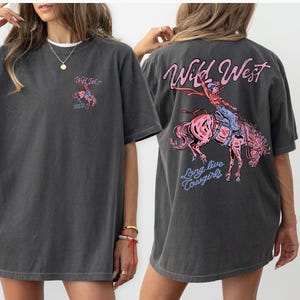 May include: Dark gray t-shirt with a graphic on the front and back. The front graphic says "Wild West" with a small image of a cowgirl riding a horse. The back graphic says "Wild West" with a cowgirl riding a horse and the words "Long live Cowgirls."
