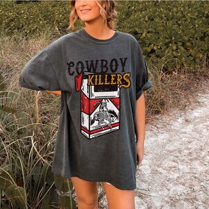 Peut inclure: Un t-shirt surdimensionn&eacute; gris fonc&eacute; avec le texte "COWBOY KILLERS" et un graphique d'un paquet de cigarettes avec un cowboy &agrave; cheval.