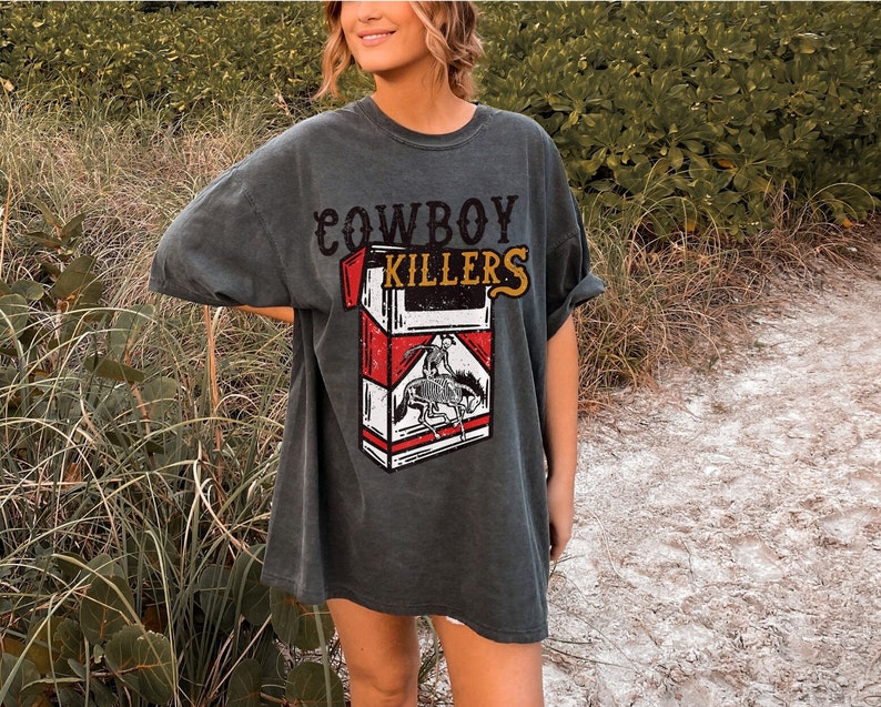 Peut inclure: Un t-shirt surdimensionn&eacute; gris fonc&eacute; avec le texte "COWBOY KILLERS" et un graphique d'un paquet de cigarettes avec un cowboy &agrave; cheval.