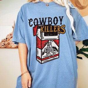 Peut inclure: Un t-shirt bleu clair avec un graphique vintage d'un paquet de cigarettes. Le paquet est rouge et blanc avec les mots "COWBOY KILLERS" imprim&eacute;s dessus. Un cow-boy squelette chevauchant un cheval est repr&eacute;sent&eacute; sur le devant du paquet.