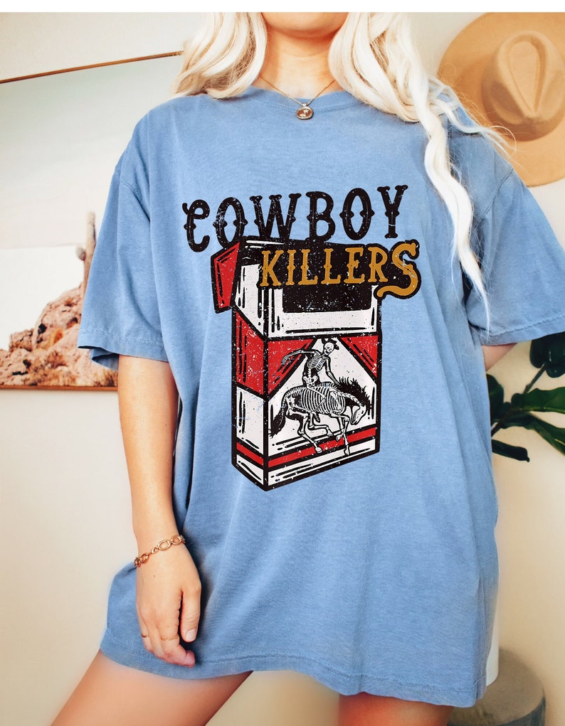 Peut inclure: Un t-shirt bleu clair avec un graphique vintage d'un paquet de cigarettes. Le paquet est rouge et blanc avec les mots "COWBOY KILLERS" imprim&eacute;s dessus. Un cow-boy squelette chevauchant un cheval est repr&eacute;sent&eacute; sur le devant du paquet.