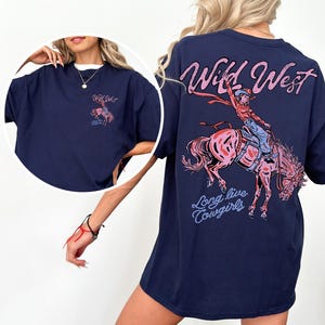 Puede incluir: Camiseta azul marino con un dise&ntilde;o de una vaquera montando un caballo salvaje, con las palabras "Wild West" y "Long live Cowgirls" en rosa y azul. La camiseta tiene un corte holgado.