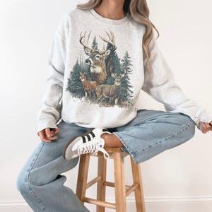 May include: A light grey sweatshirt with a detailed illustration of deer in a forest. The graphic shows a buck with large antlers, two smaller deer, and evergreen trees. The sweatshirt is worn with blue jeans.