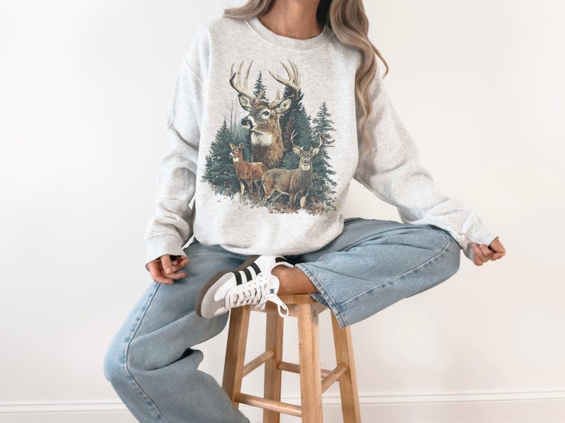 May include: A light grey sweatshirt with a detailed illustration of deer in a forest. The graphic shows a buck with large antlers, two smaller deer, and evergreen trees. The sweatshirt is worn with blue jeans.