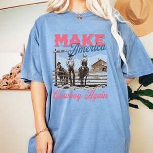 May include: A light blue t-shirt featuring a graphic print of two cowboys on horseback. The graphic includes the text "Make America Cowboy Again" and a sign that reads "Wild West".