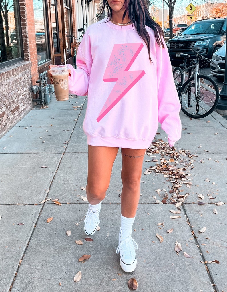 May include: A pink sweatshirt with a large, pink lightning bolt graphic. The lightning bolt has a white speckled design. The sweatshirt is worn by a person wearing white sneakers.