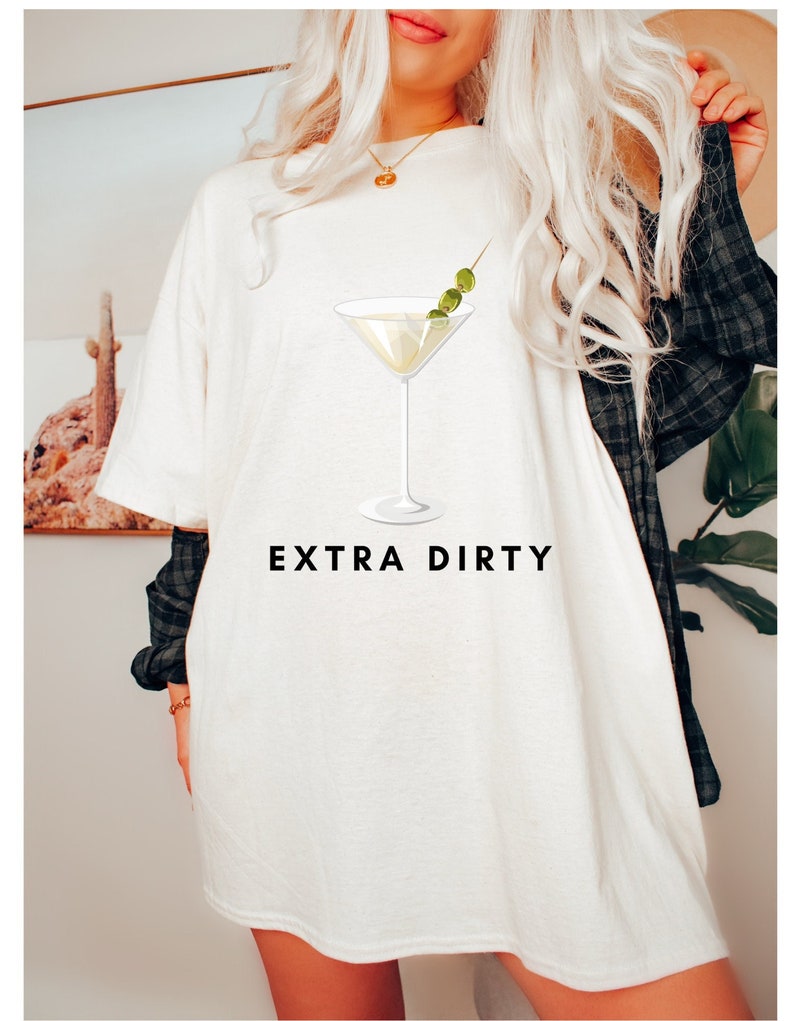 May include: White t-shirt with a martini glass graphic and the text "EXTRA DIRTY". The martini glass has a clear liquid and three green olives on a toothpick. The shirt is a casual style.