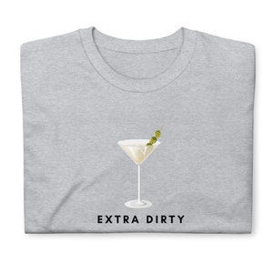 May include: A light gray t-shirt featuring a martini glass graphic with three olives on a toothpick. The words "EXTRA DIRTY" are printed in black below the glass. The shirt is folded.