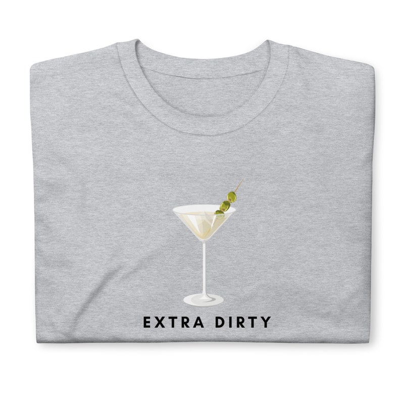May include: A light gray t-shirt featuring a martini glass graphic with three olives on a toothpick. The words "EXTRA DIRTY" are printed in black below the glass. The shirt is folded.
