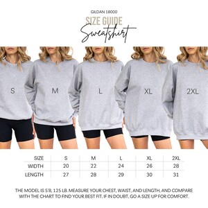 May include: Size guide for a light grey Gildan 18000 sweatshirt. The image shows models wearing sizes S, M, L, XL, and 2XL. The size chart indicates width and length in inches. The text also includes the model's height and weight.
