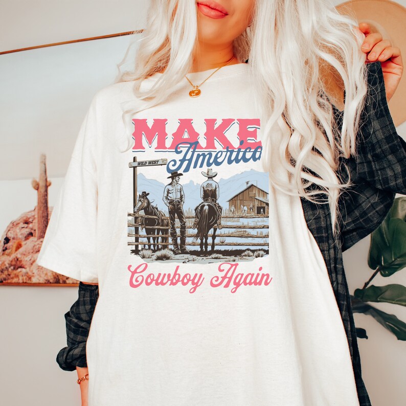 May include: White t-shirt with a vintage graphic design featuring two cowboys on horseback. The graphic includes the text "Make America Cowboy Again" with a pink and blue colour scheme.