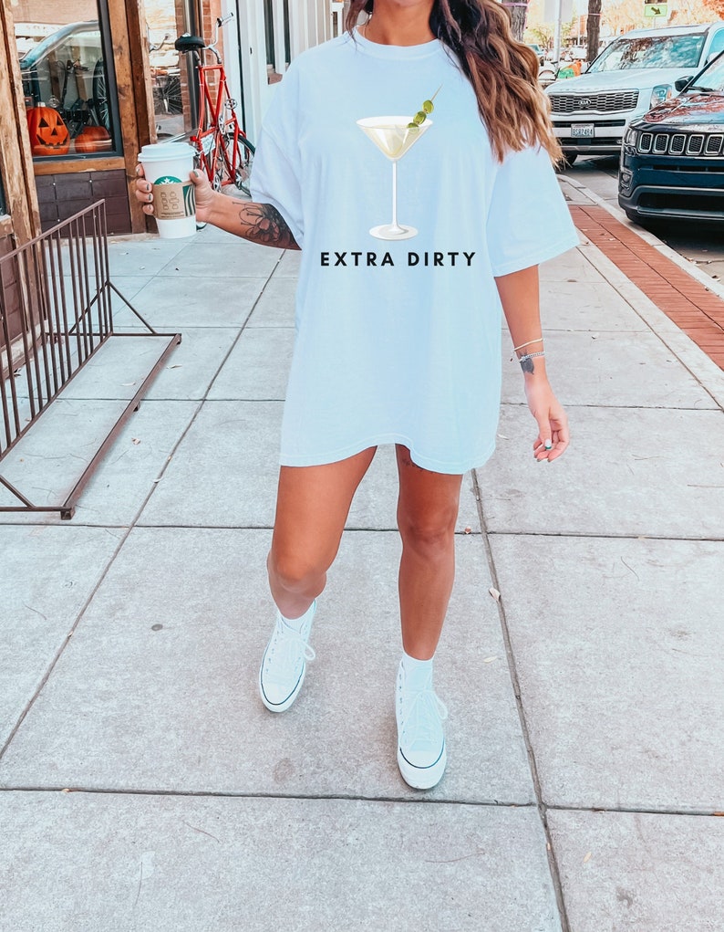 May include: White t-shirt with a martini glass graphic and the text "EXTRA DIRTY". The martini glass has a green olive garnish. The shirt is a casual, oversized style.