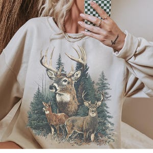 May include: Beige sweatshirt with a detailed graphic of three deer in a forest. The deer have large antlers and are surrounded by evergreen trees. The sweatshirt has long sleeves and a crew neck.