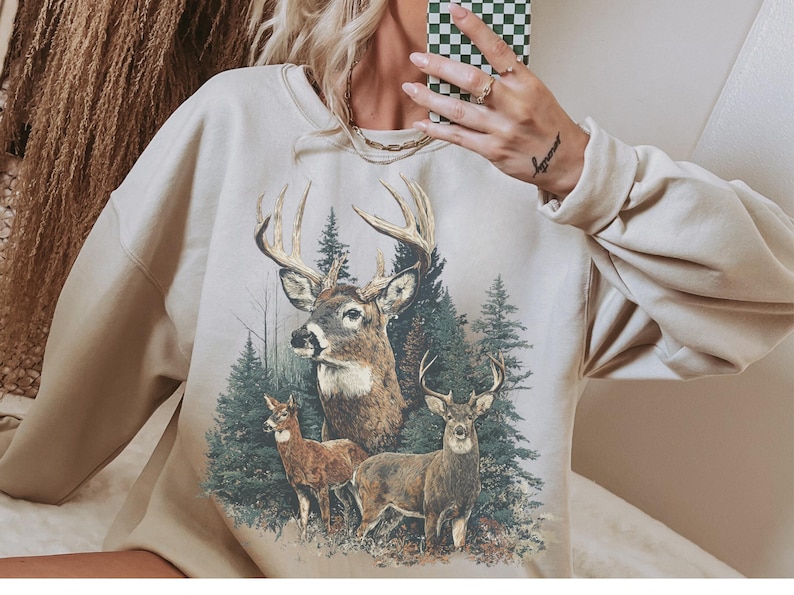 May include: Beige sweatshirt with a detailed graphic of three deer in a forest. The deer have large antlers and are surrounded by evergreen trees. The sweatshirt has long sleeves and a crew neck.