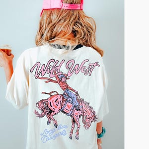 Puede incluir: Camiseta color crema con un gr&aacute;fico rosa de una vaquera montando un caballo rosa, con las palabras "Wild West" y "Long live Cowgirls" en escritura rosa y azul. La persona de la imagen lleva una gorra rosa.
