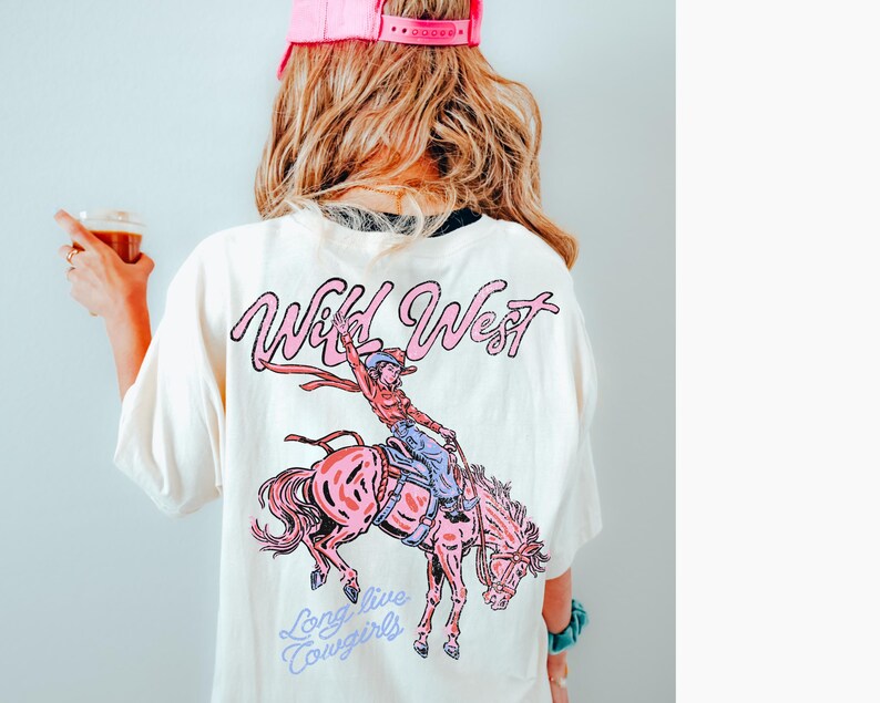 Puede incluir: Camiseta color crema con un gr&aacute;fico rosa de una vaquera montando un caballo rosa, con las palabras "Wild West" y "Long live Cowgirls" en escritura rosa y azul. La persona de la imagen lleva una gorra rosa.