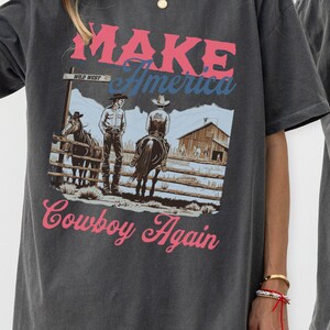 May include: A dark grey t-shirt with a graphic print of two cowboys on horseback. The graphic includes the text "MAKE America Cowboy Again" and a sign that reads "WILD WEST".