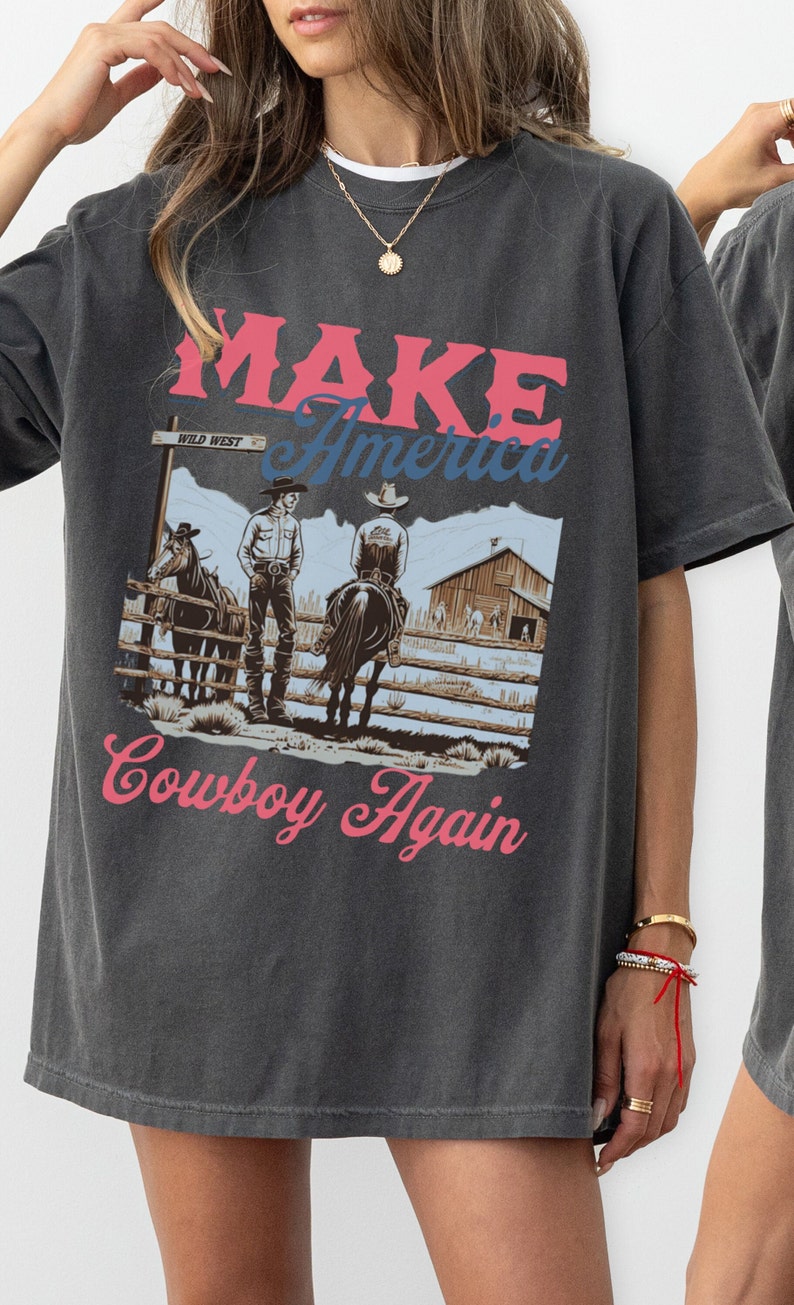 May include: A dark grey t-shirt with a graphic print of two cowboys on horseback. The graphic includes the text "MAKE America Cowboy Again" and a sign that reads "WILD WEST".