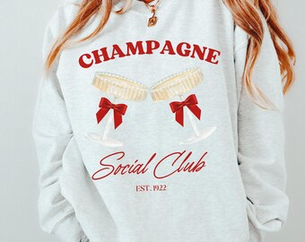 Pode incluir: Uma camisa de moletom branca com o texto "Champagne Social Club Est. 1922" em letras vermelhas. O design apresenta duas taças de champanhe com laços vermelhos.
