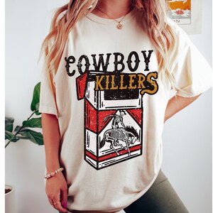 Peut inclure: Un t-shirt cr&egrave;me avec un graphique vintage d'un paquet de cigarettes qui dit "Cowboy Killers". Le graphique repr&eacute;sente un squelette &agrave; cheval.