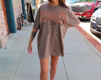 May include: Brown oversized t-shirt with the word "COWGIRL" in coral lettering and a barbed wire design. The shirt is a relaxed fit and appears to be made of a soft material. The shirt is worn outdoors.