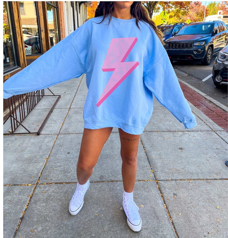 May include: A light blue sweatshirt with a pink lightning bolt graphic. The sweatshirt has long sleeves and a crew neck.
