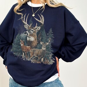 May include: Navy blue sweatshirt with a detailed illustration of deer in a forest. The design features three deer of different sizes and evergreen trees. The sweatshirt has a crew neck and long sleeves.