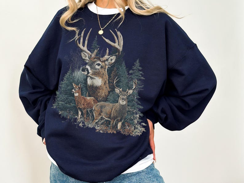 May include: Navy blue sweatshirt with a detailed illustration of deer in a forest. The design features three deer of different sizes and evergreen trees. The sweatshirt has a crew neck and long sleeves.