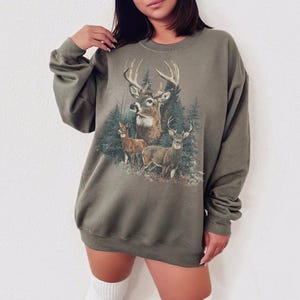 May include: Khaki green sweatshirt with a detailed graphic of deer in a forest setting. The design includes a large buck with prominent antlers, two smaller deer, and evergreen trees. The sweatshirt has long sleeves and a relaxed fit.