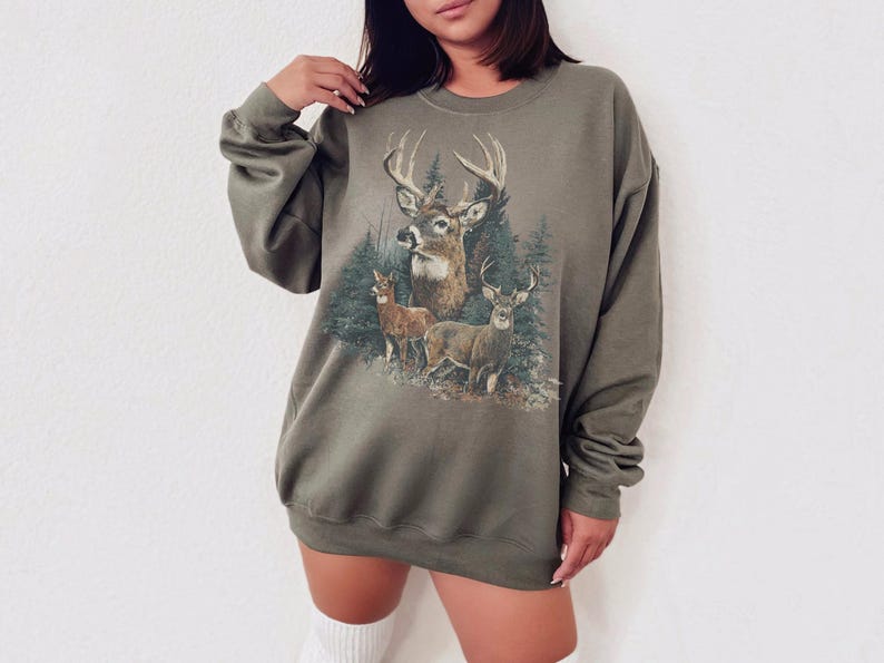 May include: Khaki green sweatshirt with a detailed graphic of deer in a forest setting. The design includes a large buck with prominent antlers, two smaller deer, and evergreen trees. The sweatshirt has long sleeves and a relaxed fit.